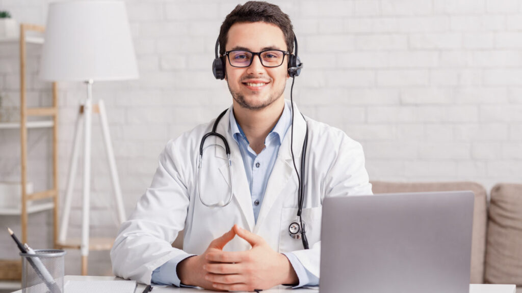 Health blog. Young doctor in headphones sitting at table with laptop